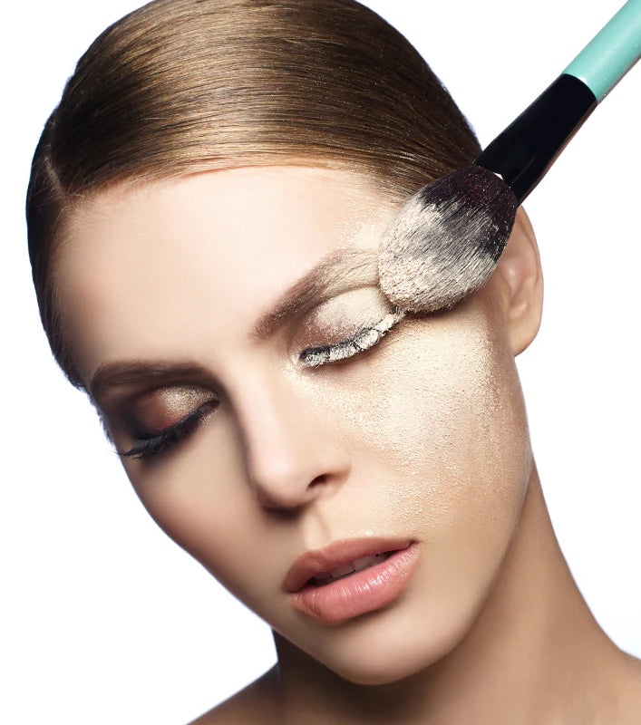 A woman with closed eyes, applying powder to her face using a makeup brush.
