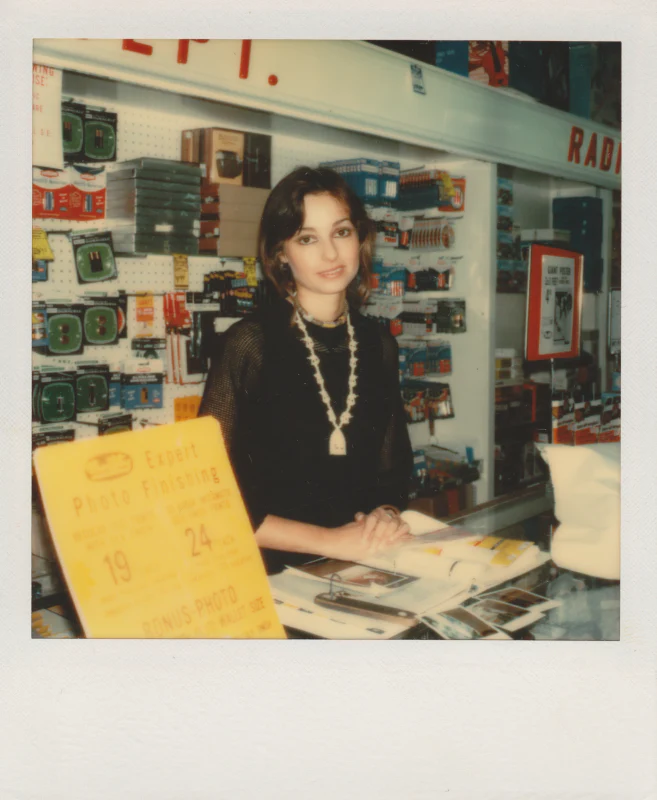 Vintage Polaroid of a woman behind a store counter, wearing a statement necklace, surrounded by various products.