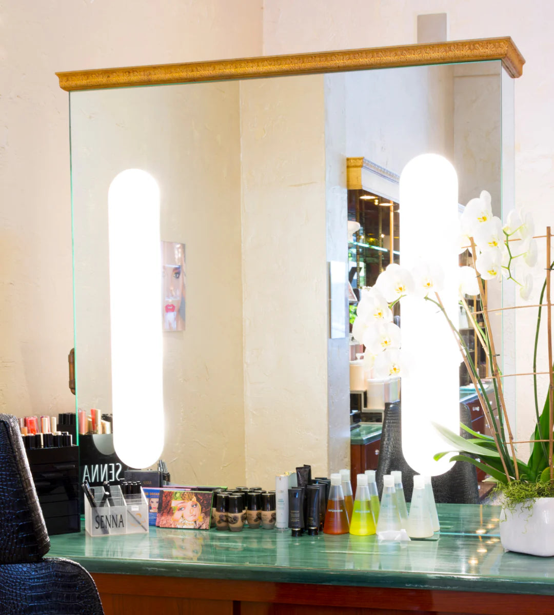 A vanity mirror with lights, makeup products, and orchids on a glass table.