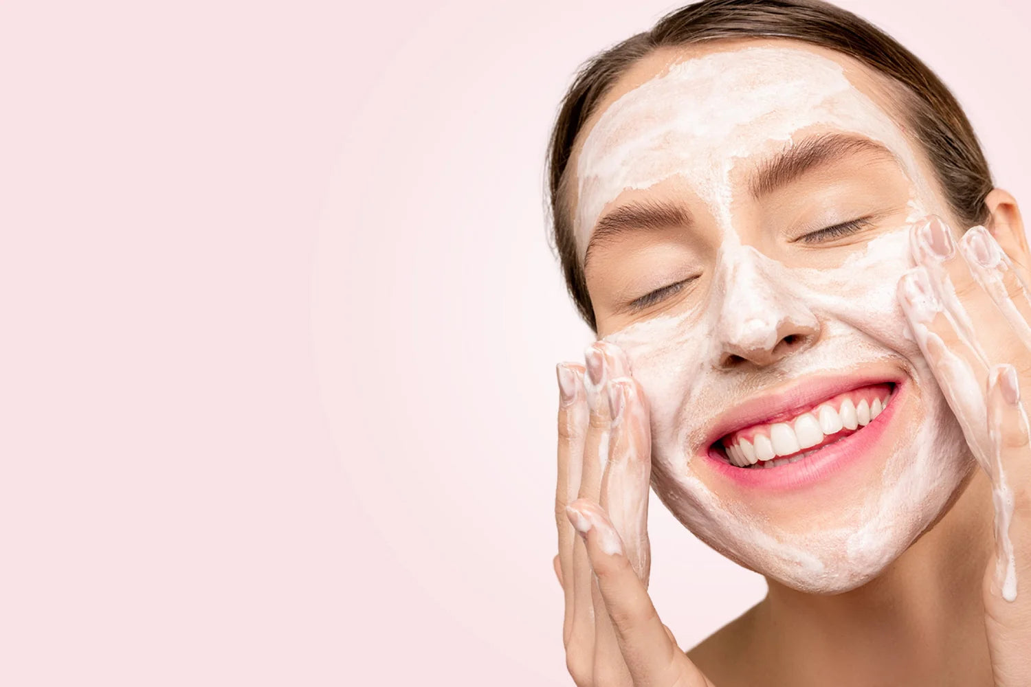 A smiling woman applying a foaming facial cleanser on her face against a pink background.