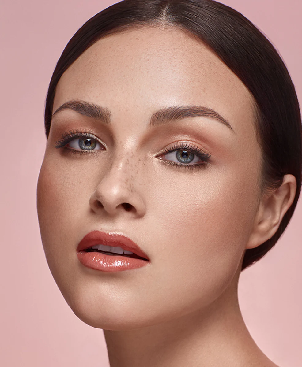 A model with a soft pink makeup look, glossy lips, and subtle freckles on a pink background.